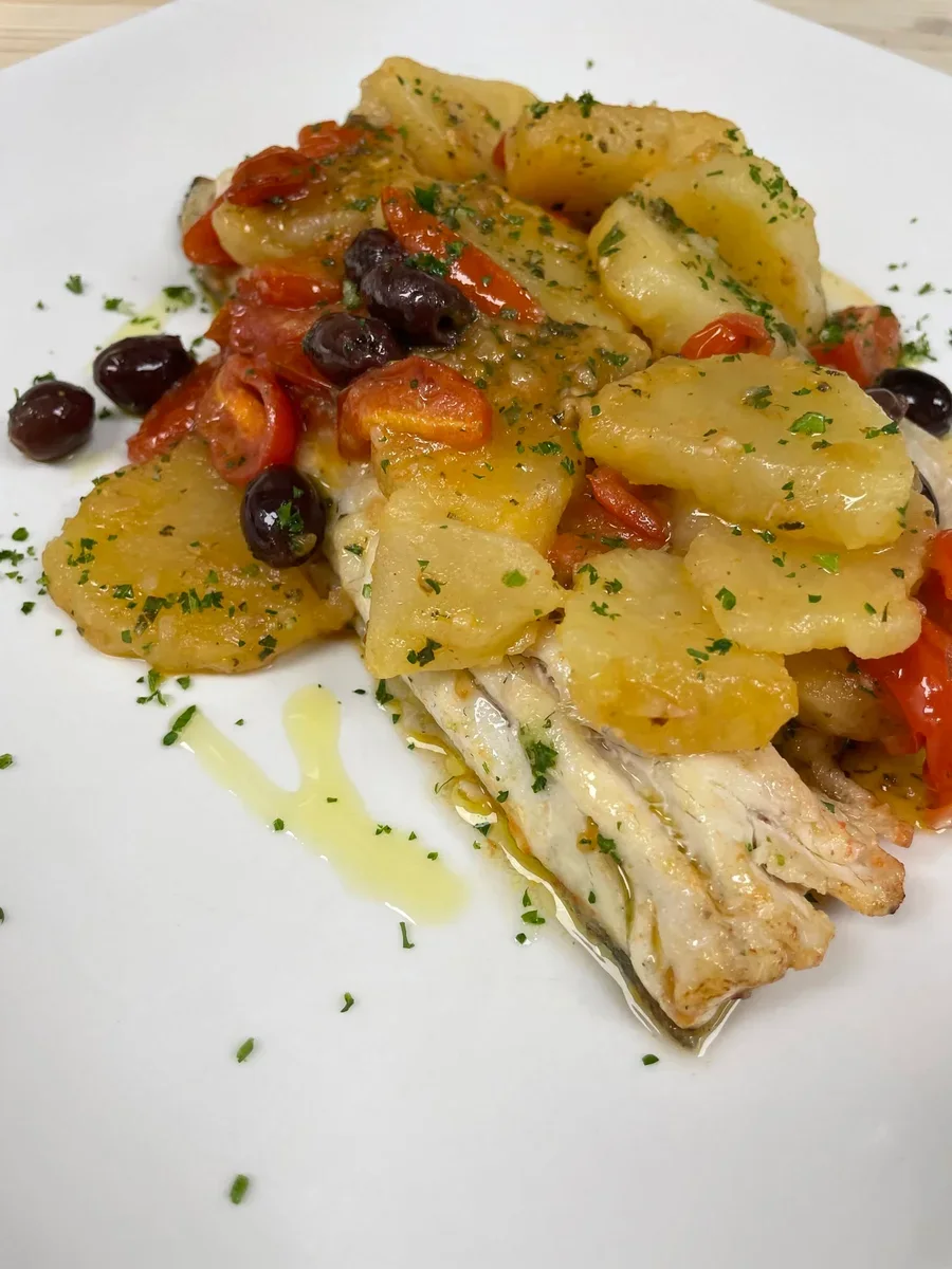 Sea bass fillet with sliced potatoes, grape tomatoes, black olives and chopped herbs on a white plate